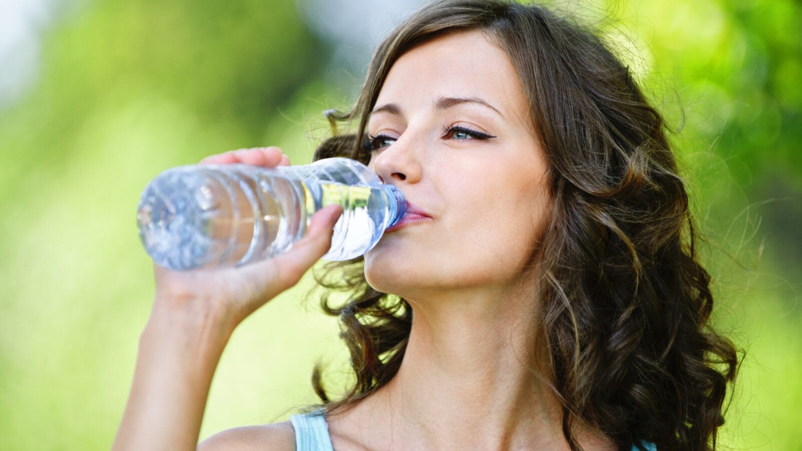 Portrait of young beautiful dark-haired woman wearing blue t-shirt drinking water at summer green park.
