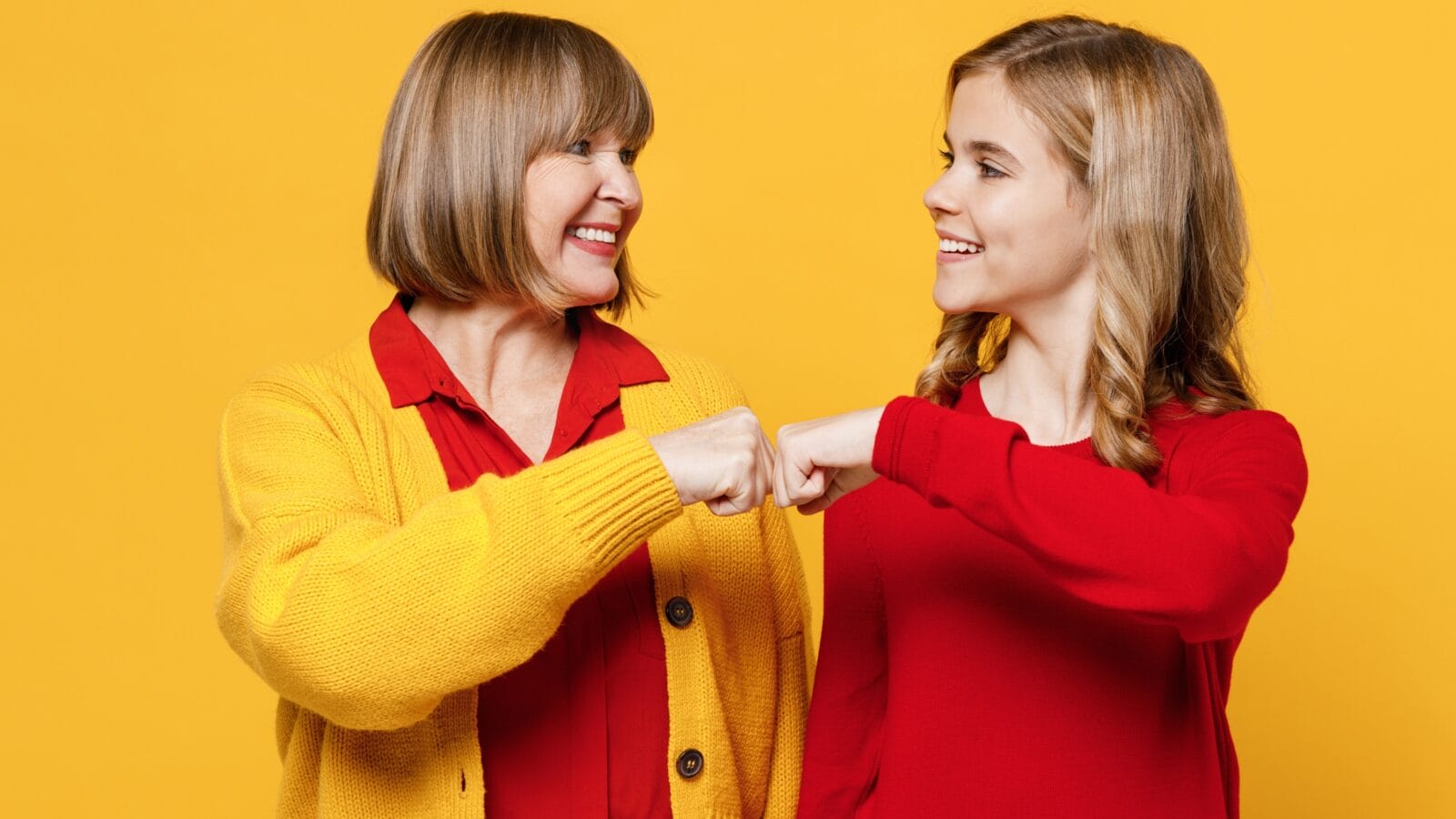 Grandmother granddaughter do fist bump gesture look to each other isolated on plain yellow background.