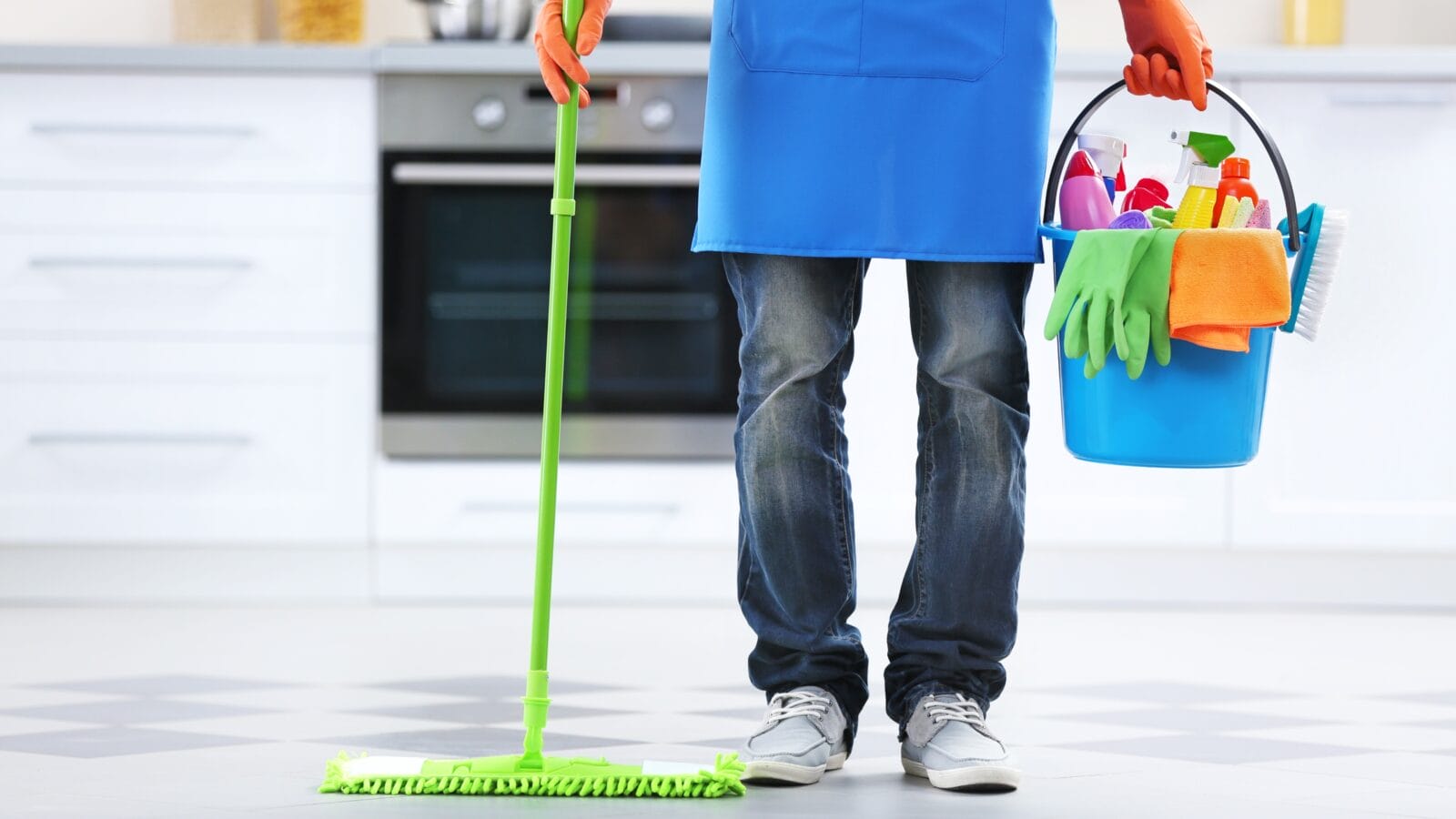 Man holding mop and plastic bucket with brushes, gloves and detergents in the kitchen.