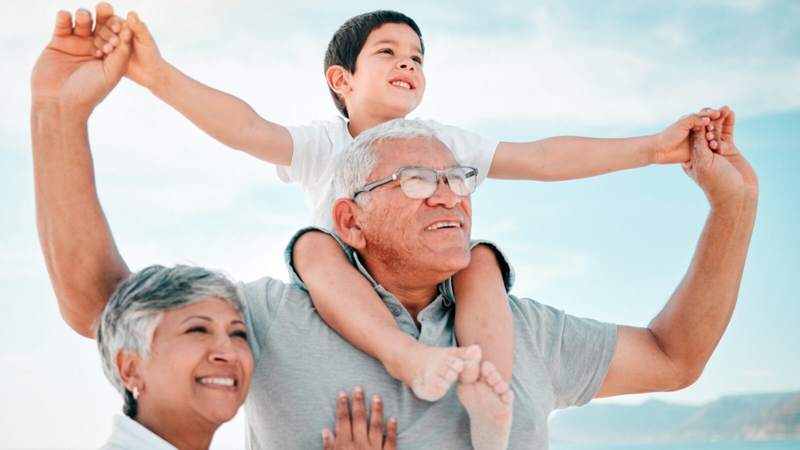 A kid on grandfathers shoulder with grandmother beside them walking on a beach.