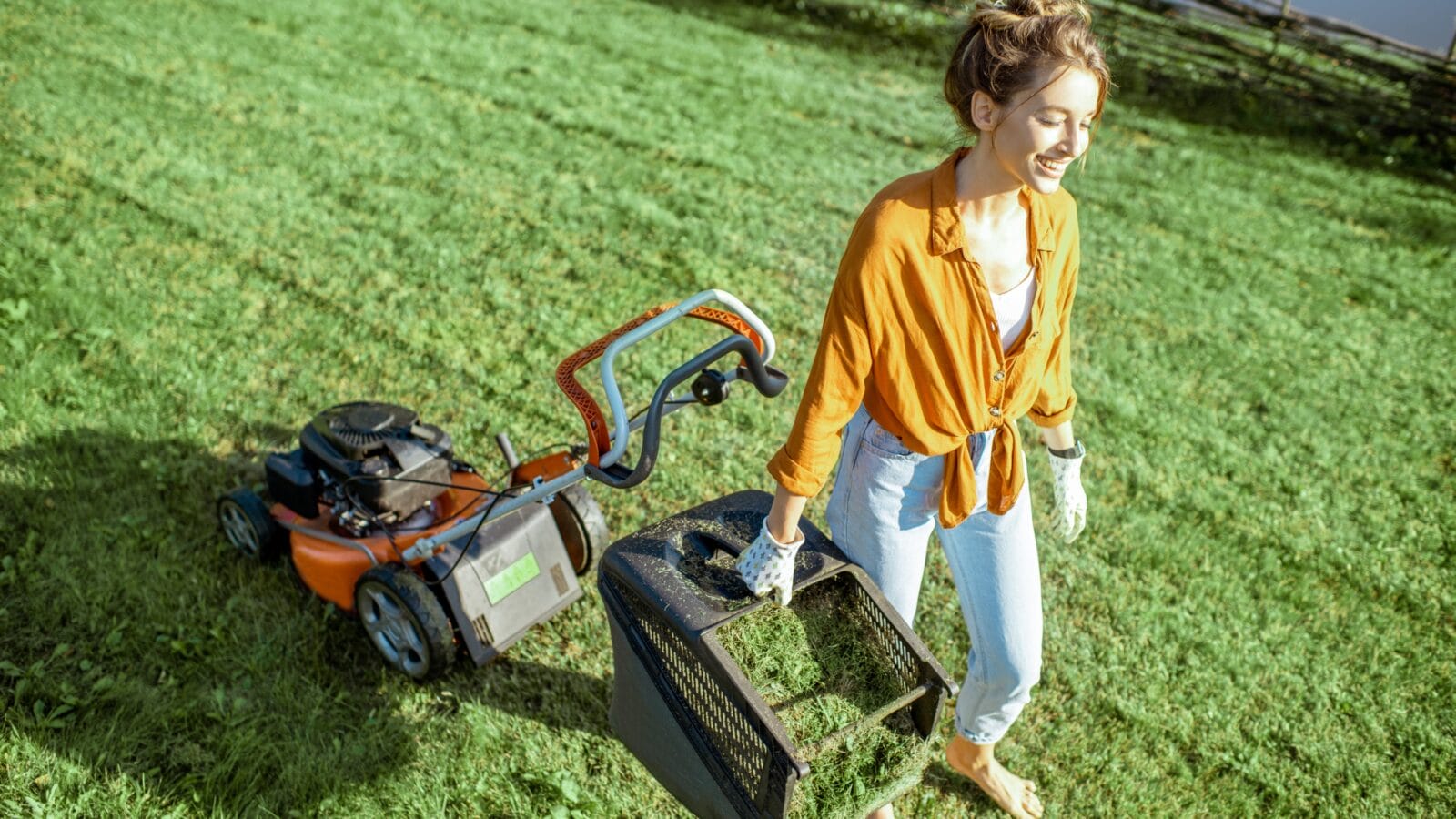 Beautiful young woman carrying basket full of cut grass and a lawn mower behind.