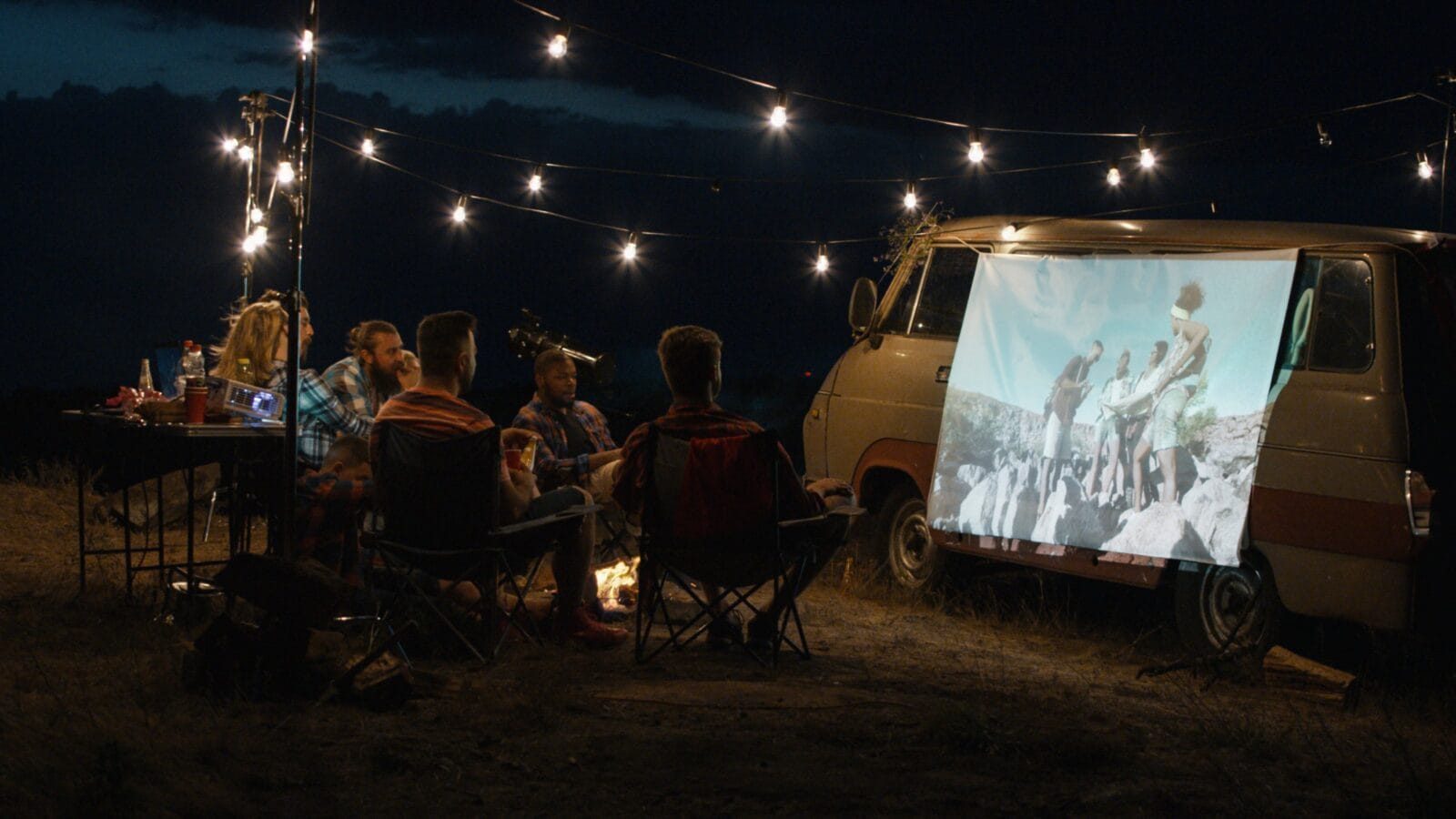 Friends gathering in campsite around bonfire and watching movie with projector on van side in dark evening