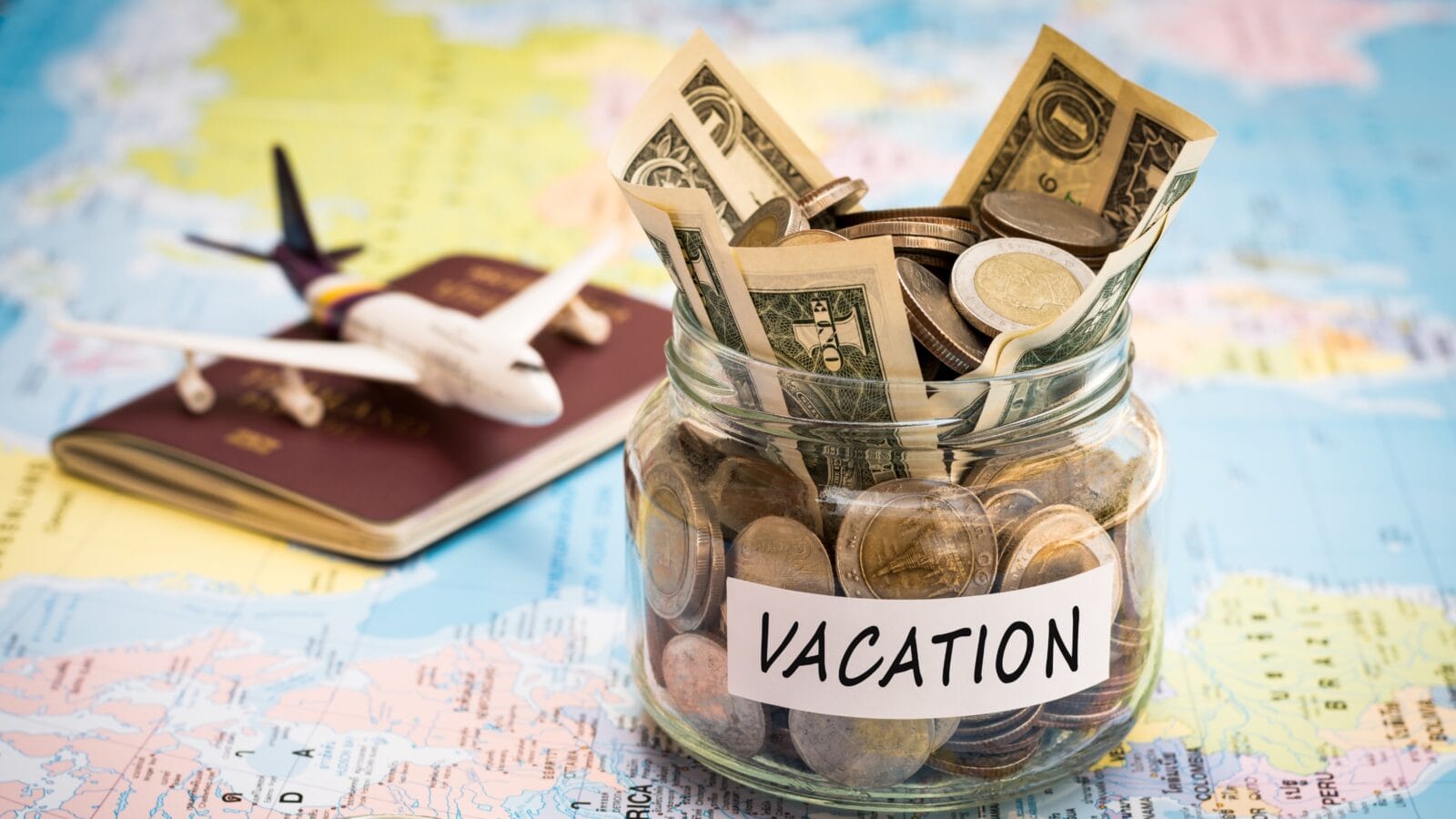 Vacation money savings in a glass jar with passport and aircraft toy on world map.