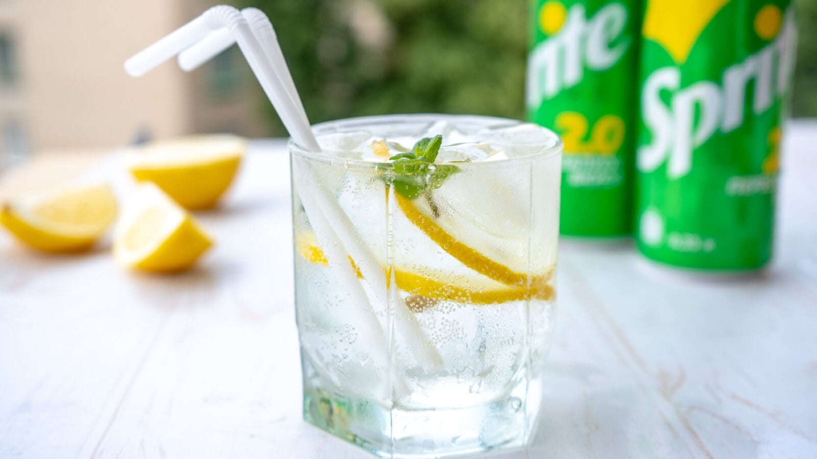 A glass of soda with ice and lemon, with sprite cans in the background.