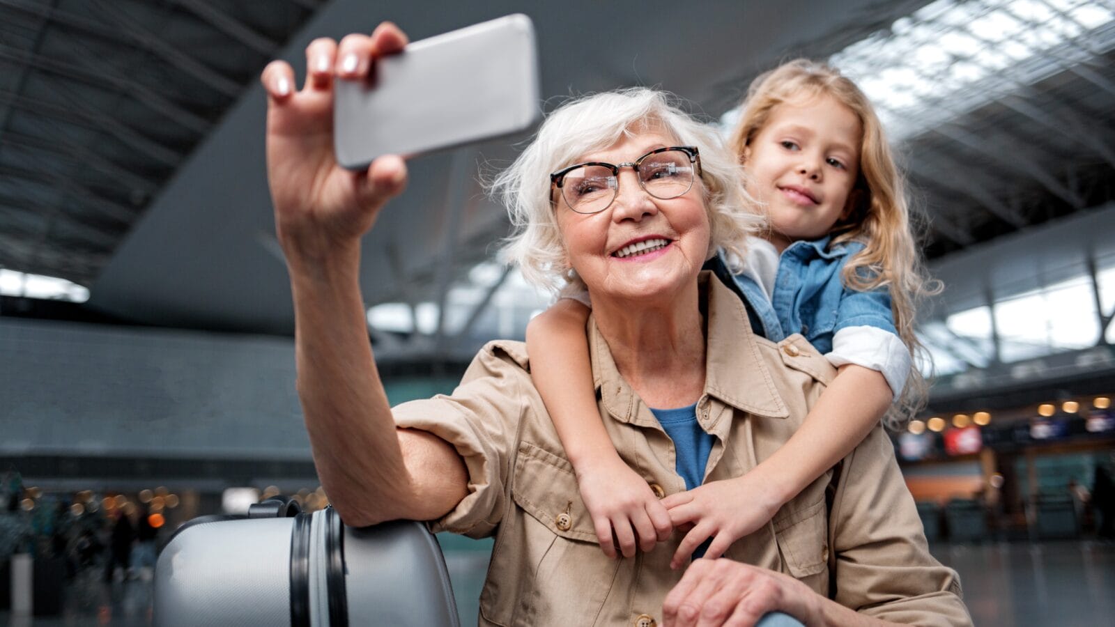 Portrait of cheerful aged charming grandmother is standing at airport lounge with her little granddaughter.