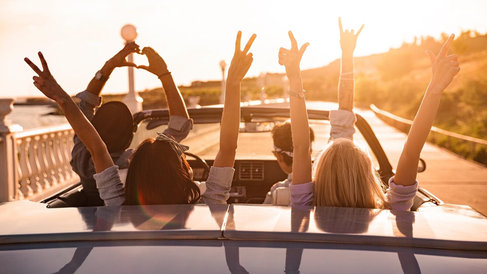 Group of happy young friends in cabriolet with raised hands driving on sunset