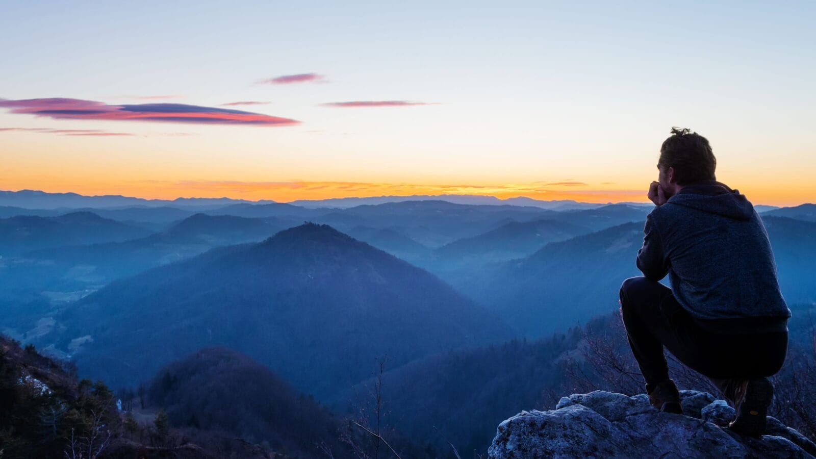 Male hiker crouching on top of the hill and enjoying scenic view of twilight landscape below