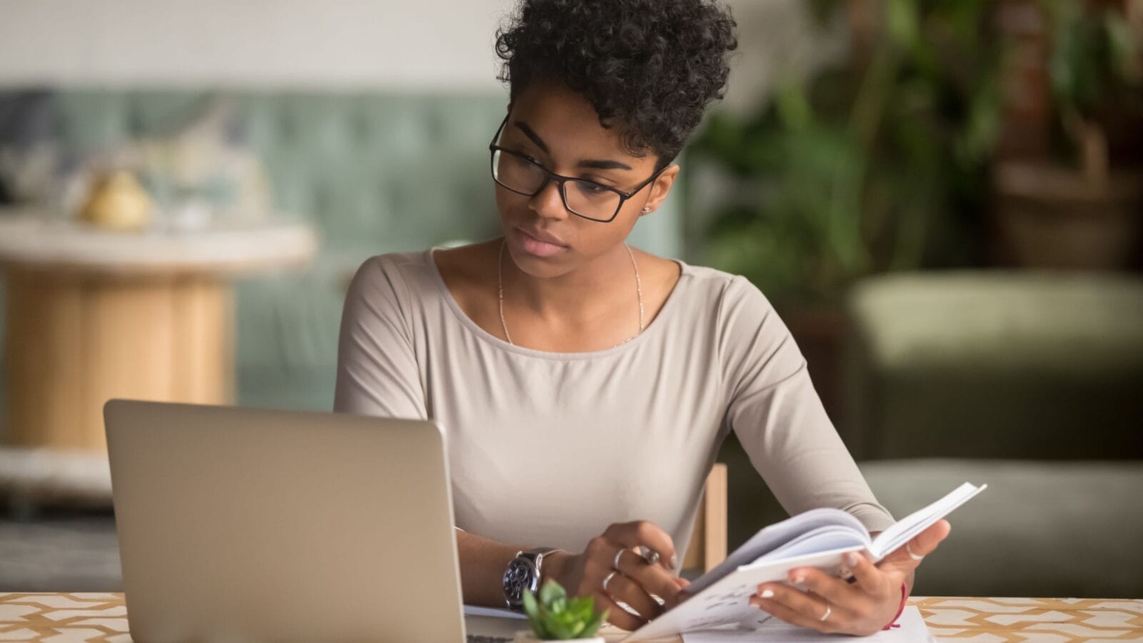 Focused young african american businesswoman or student looking at laptop holding book learning, serious black woman working or studying with computer doing research or preparing for exam online