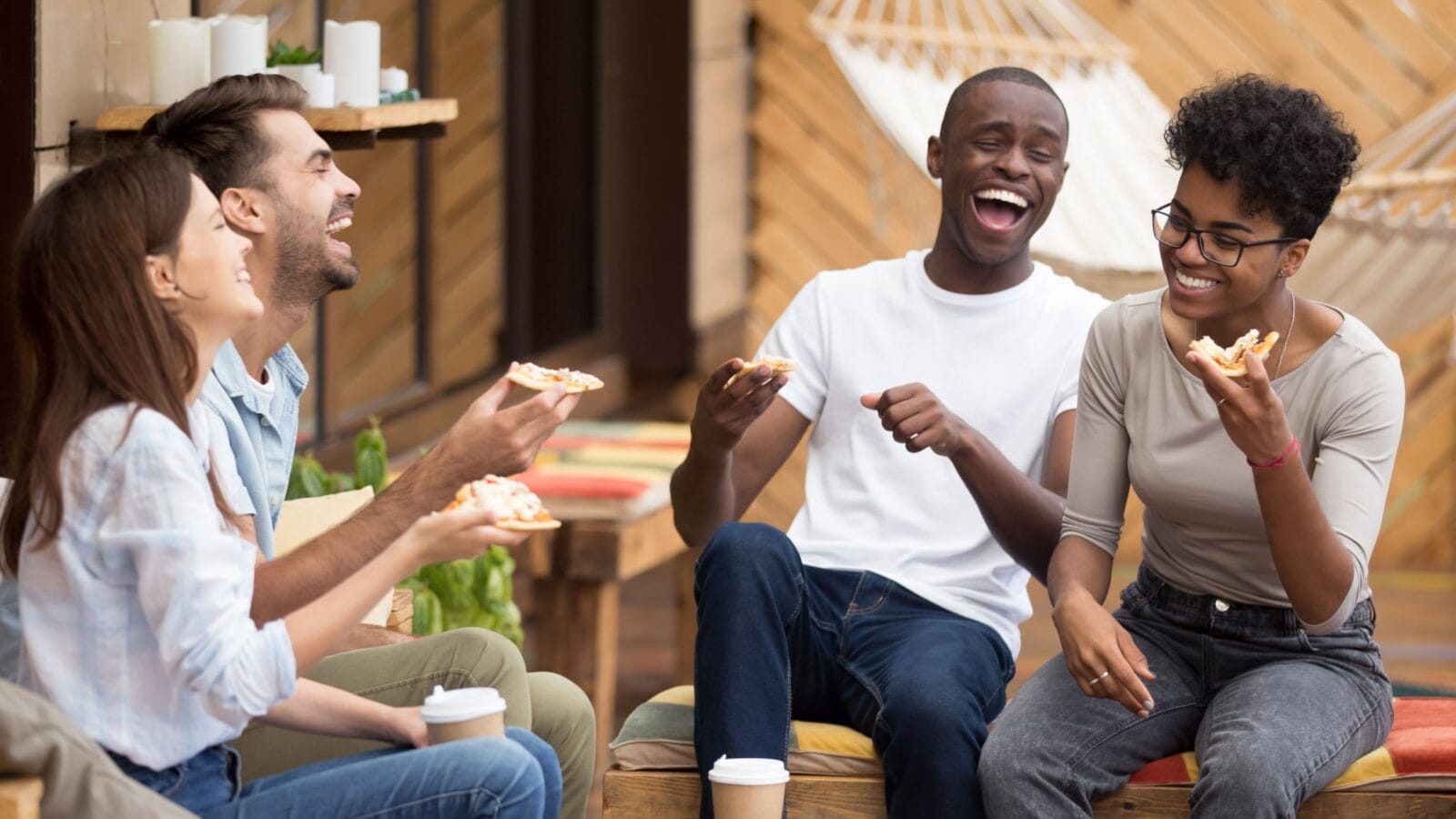 Excited multiethnic millennial friends sit in restaurant outside eat delicious pizza laugh having fun