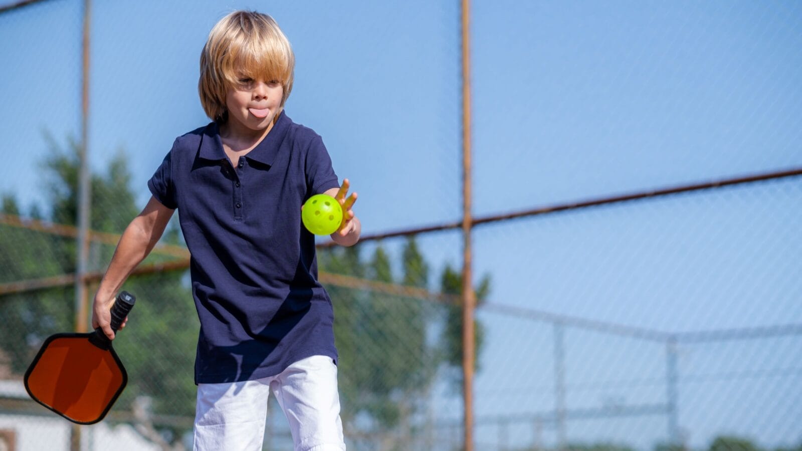 Happy blonde boy playing pickleball game, hitting pickleball yellow ball with paddle, outdoor sport leisure kids activity