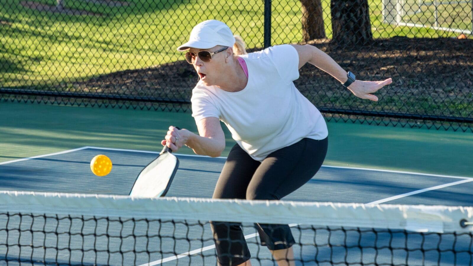 A female pickleball player returns a volley of a bright yellow ball at the net on a dedicated court at a public park
