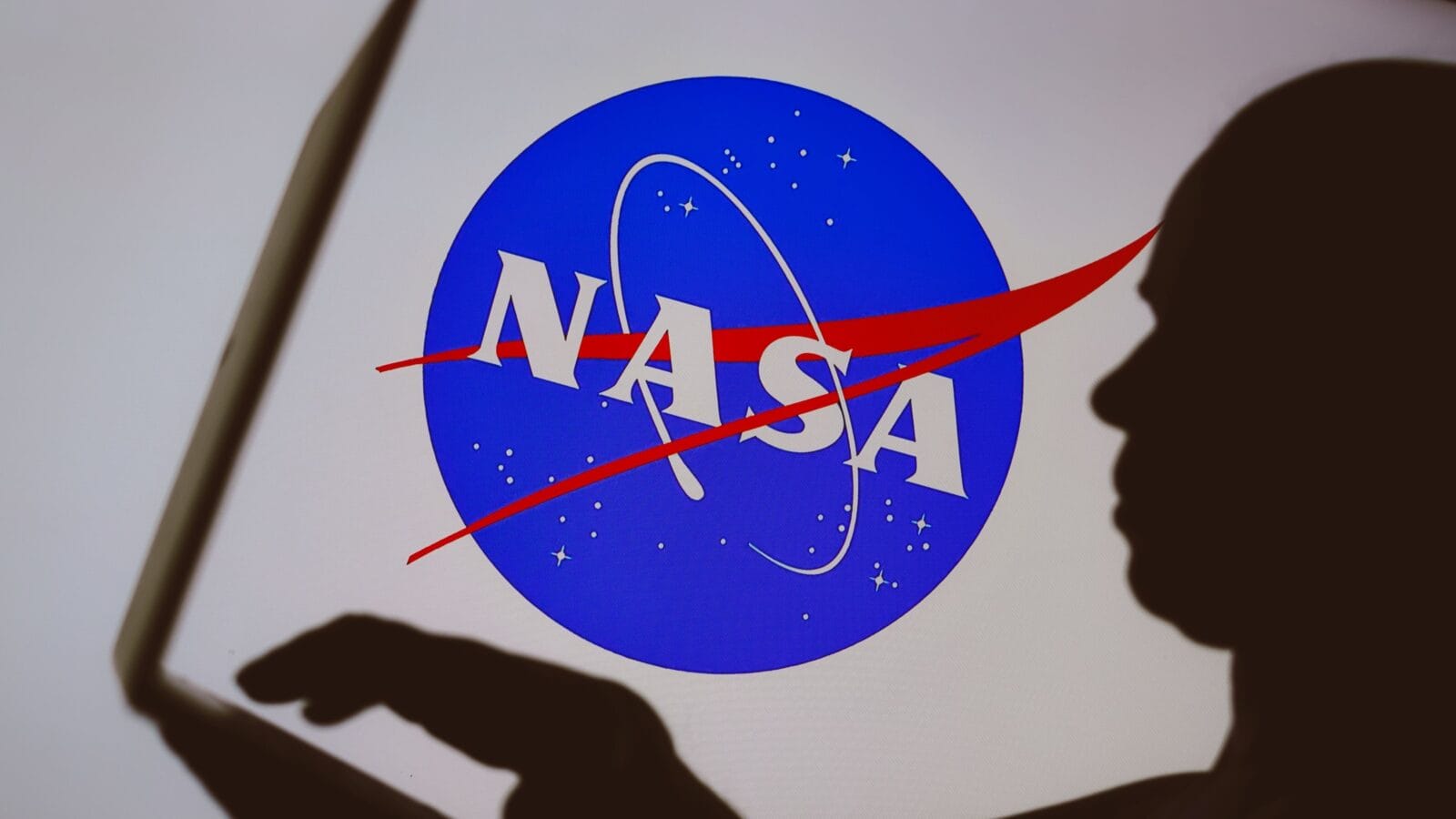 In this photo illustration, the National Aeronautics and Space Administration (NASA) logo is seen in the background of a silhouette of a person using a notebook
