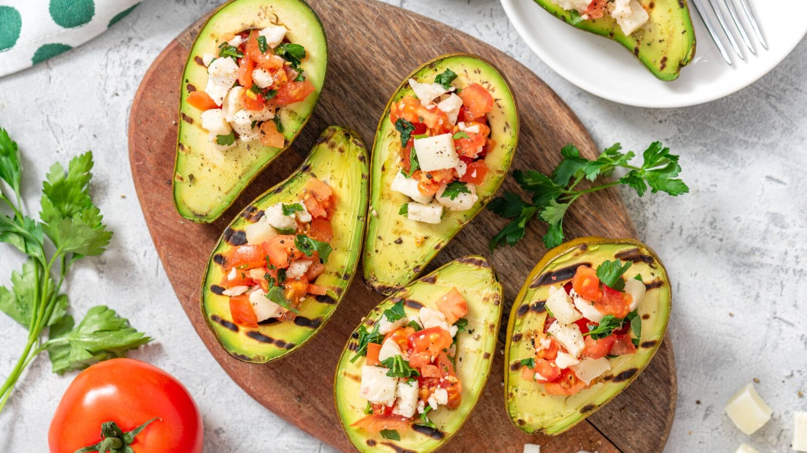Grilled avocado stuffed with tomatoes, goat cheese, parsley, and olive oil on a cutting board.