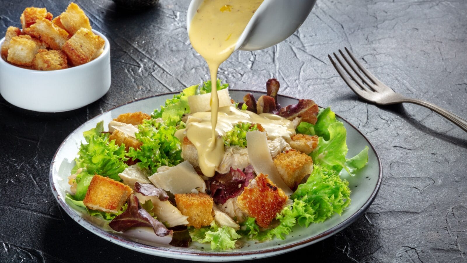 Chicken Caesar salad with the classic dressing being poured, croutons, and pepper