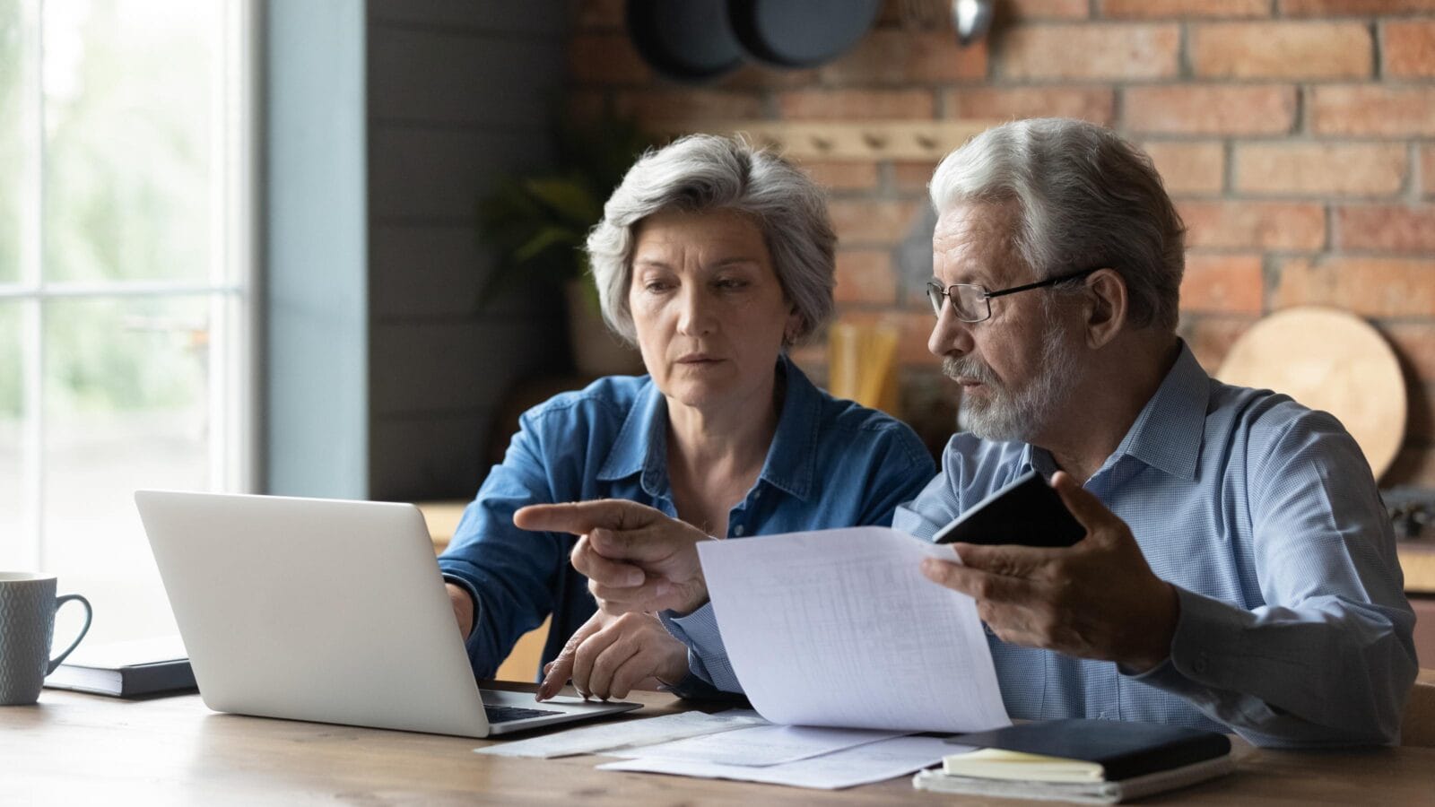 Focused senior husband and wife sit at table at home look at laptop screen pay bills taxes online.
