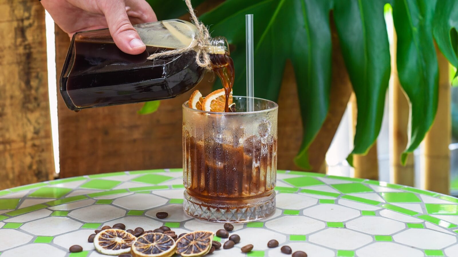 Hand pouring cold brew coffee in a bottle with transparent glass full of ice on green leaves background