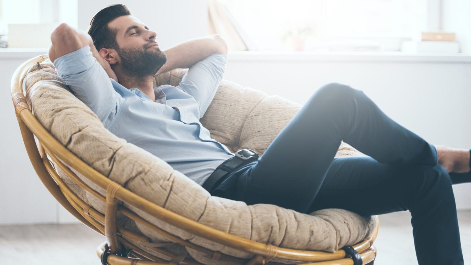 Handsome young man keeping eyes closed and holding hands behind head while sitting in big comfortable chair at home