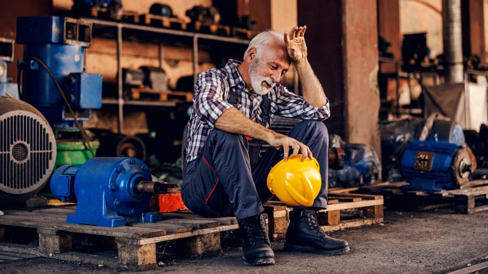 An old, tired bearded factory worker in overalls is sitting on the pallet and taking a break from hard work.
