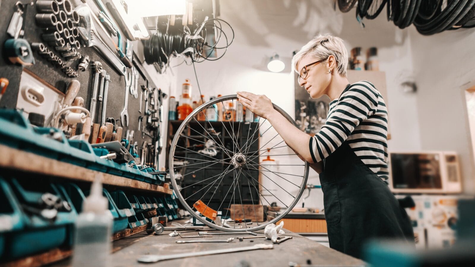 Cute Caucasian female worker holding and repairing bicycle wheel while standing in bicycle workshop.