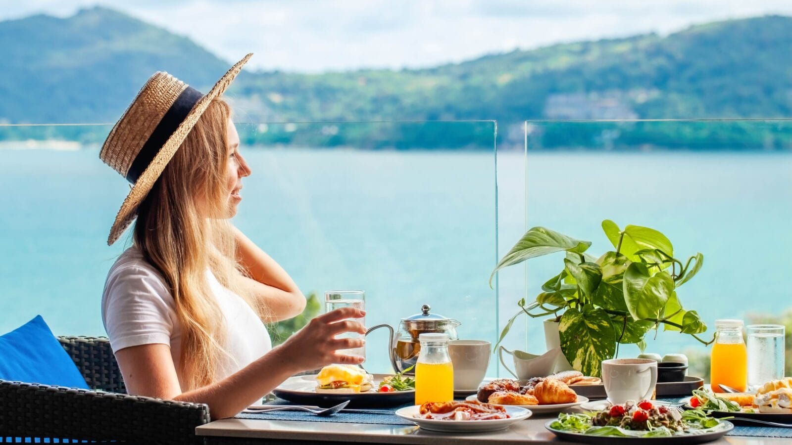 Smiling woman sitting at table in cafe with sea view drinking glass of orange juice, Phuket island, Thailand, Asia