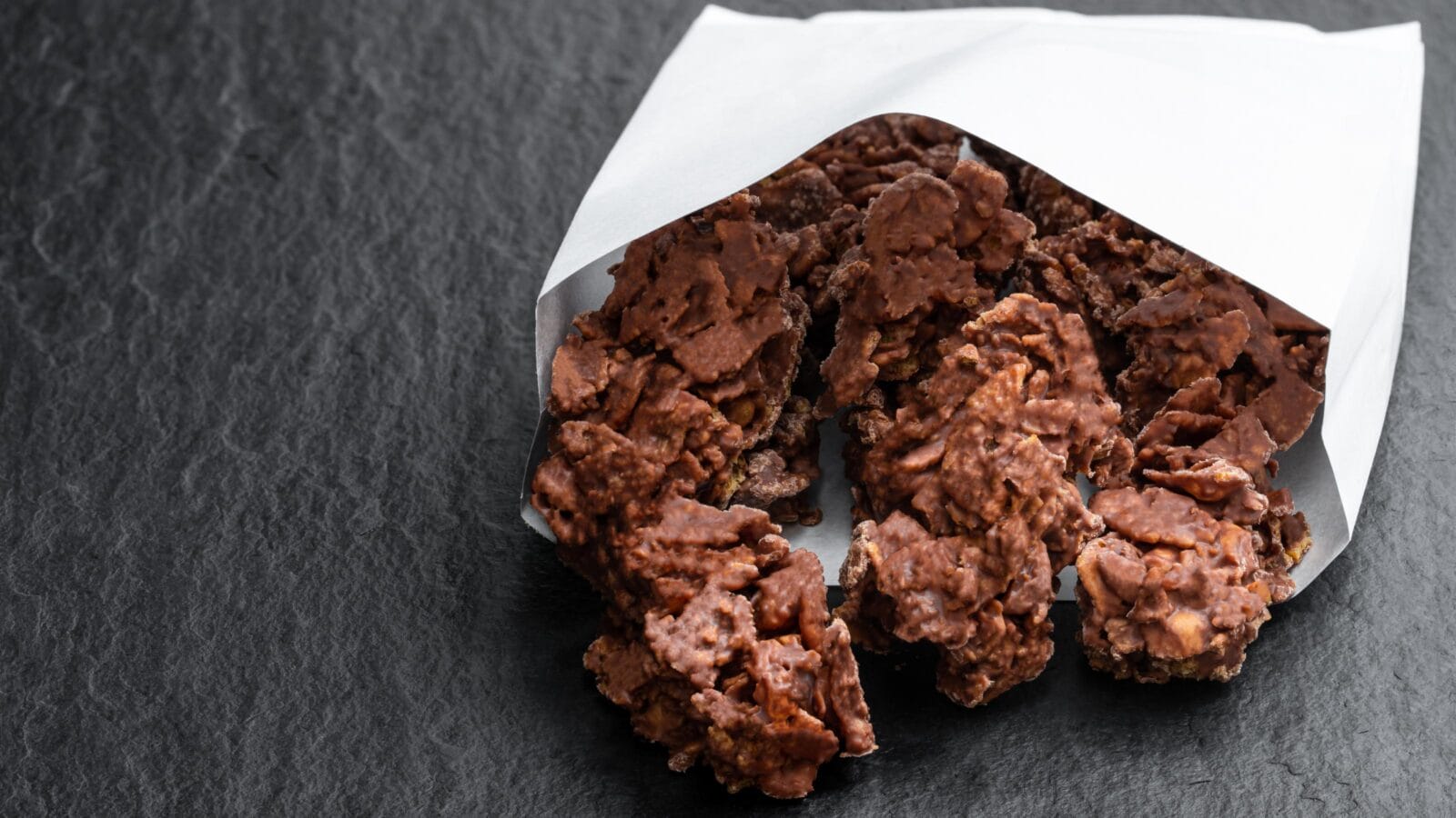 Chocolate corn flake clusters in paper bag on black stone background