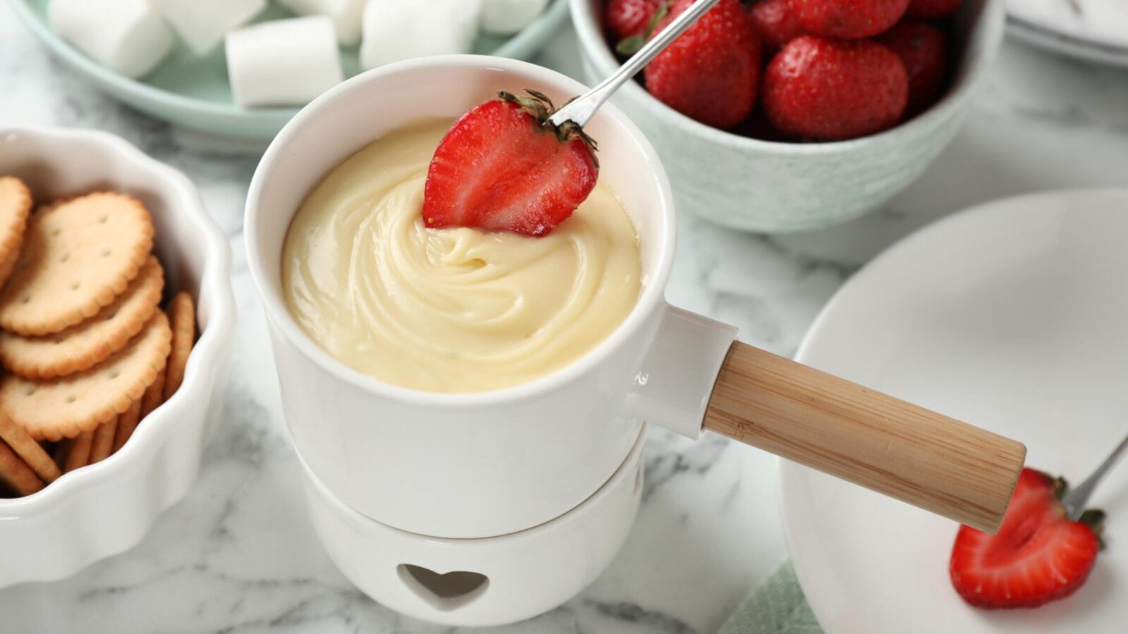 Dipping strawberry into fondue pot with white chocolate on marble table