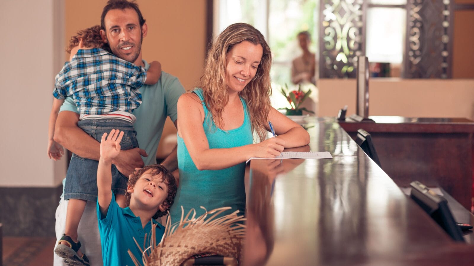 Middle-aged woman checking in, while her husband carrying a tired child, and her other son waving at the camera