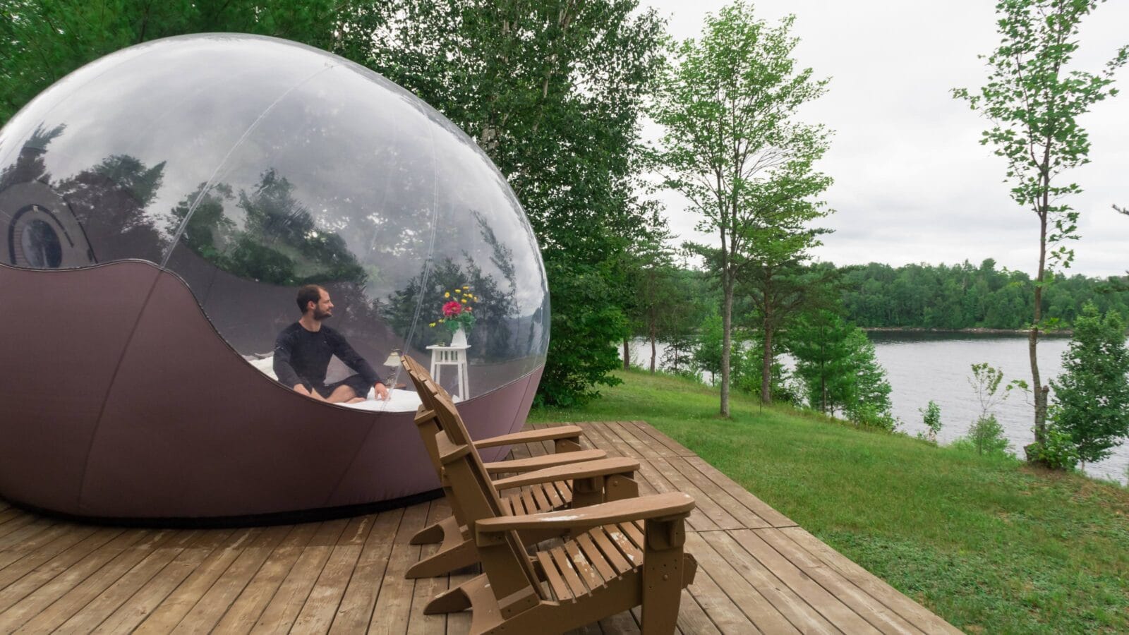 man sitting on a bed inside a transparent bubble tent facing a lake