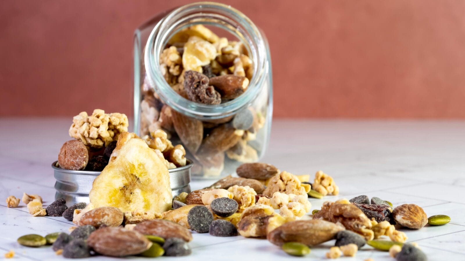 Glass jar of nuts, trail mix, granola, banana chips, dried fruit, chocolate chips, almonds, peanuts, walnuts and sunflower seeds.