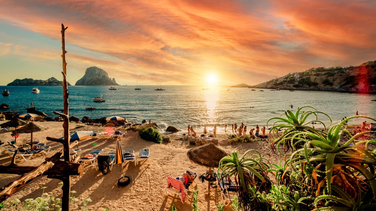 Picturesque view of Cala d'Hort tropical Beach, people hangout in beautiful beach with Es Vedra rock view during magnificent vibrant sunset glowing sun.