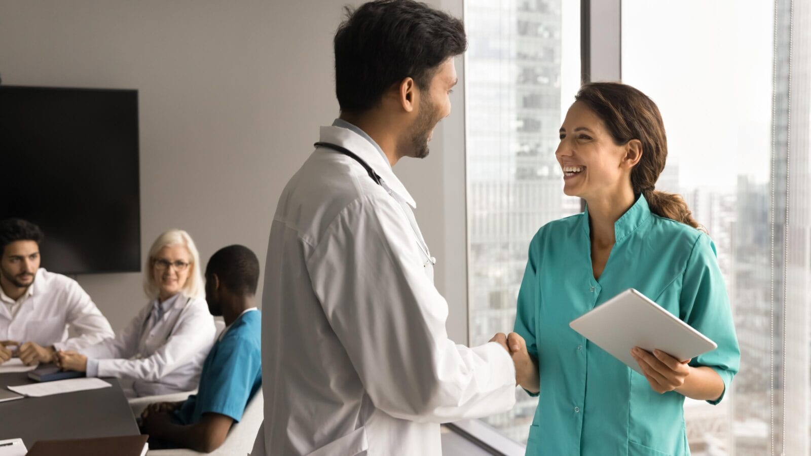 Positive young medical colleagues shaking hands in meeting room with team talking in background.