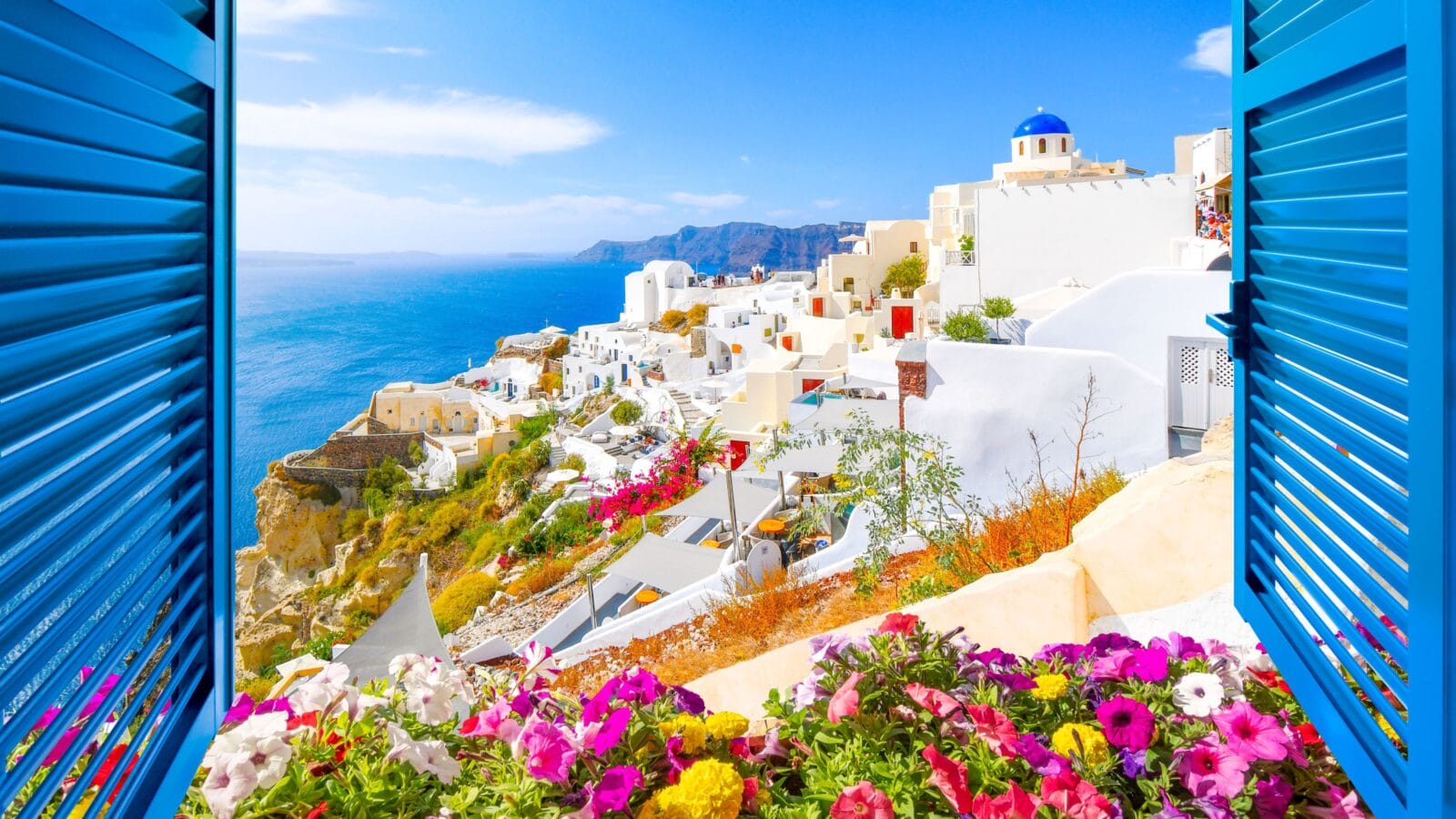 Hillside view through an open window with blue shutters of the blue dome church, caldera, sea and white village of Oia on the island of Santorini, Greece.