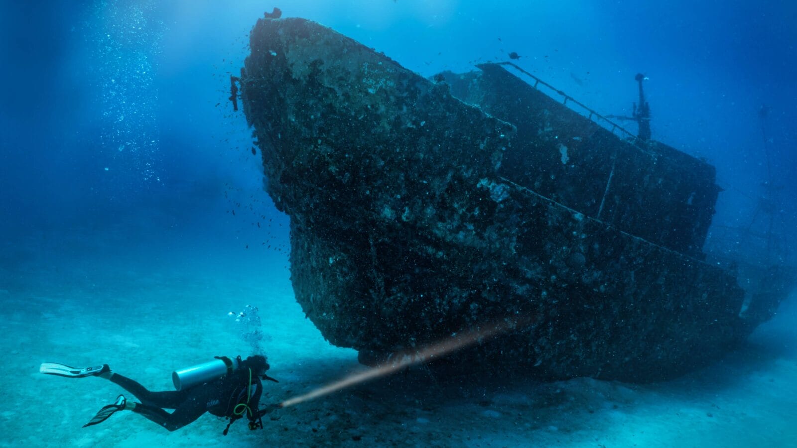 A scuba diver with a torch explores a sunken shipwreck at the seabed of the Maldives islands, Indian Ocean