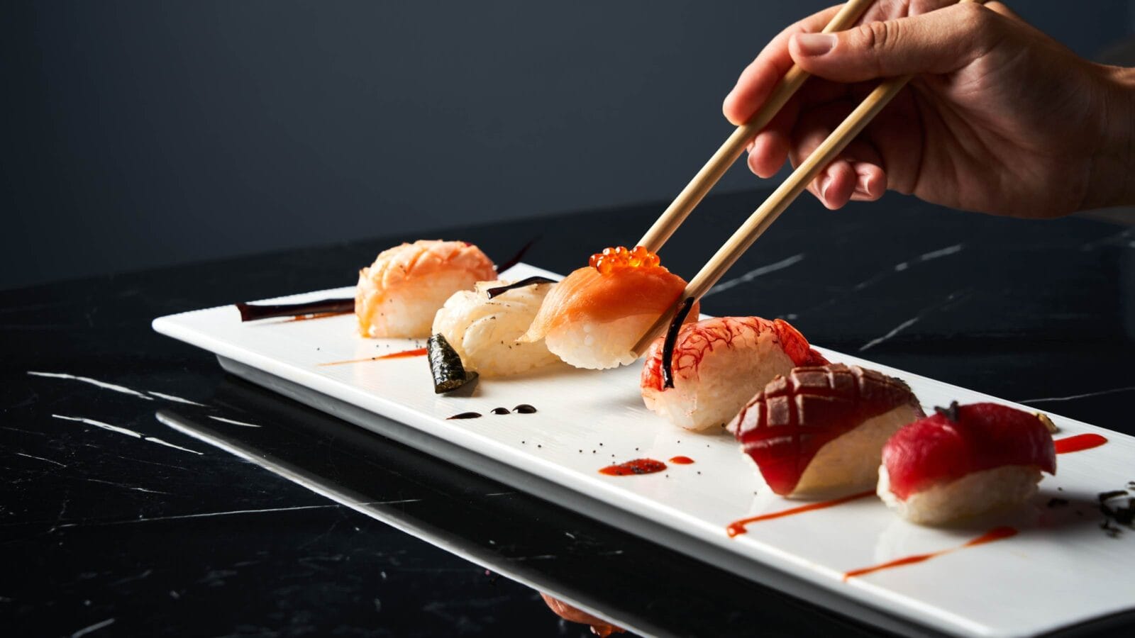 Hand holding bamboo chopsticks an elegant piece of sushi while soaking it in soy sauce at restaurant.